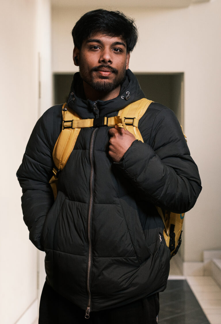 A delivery worker in a black padded jacket and yellow backpack holds a shoulder strap in a doorway.