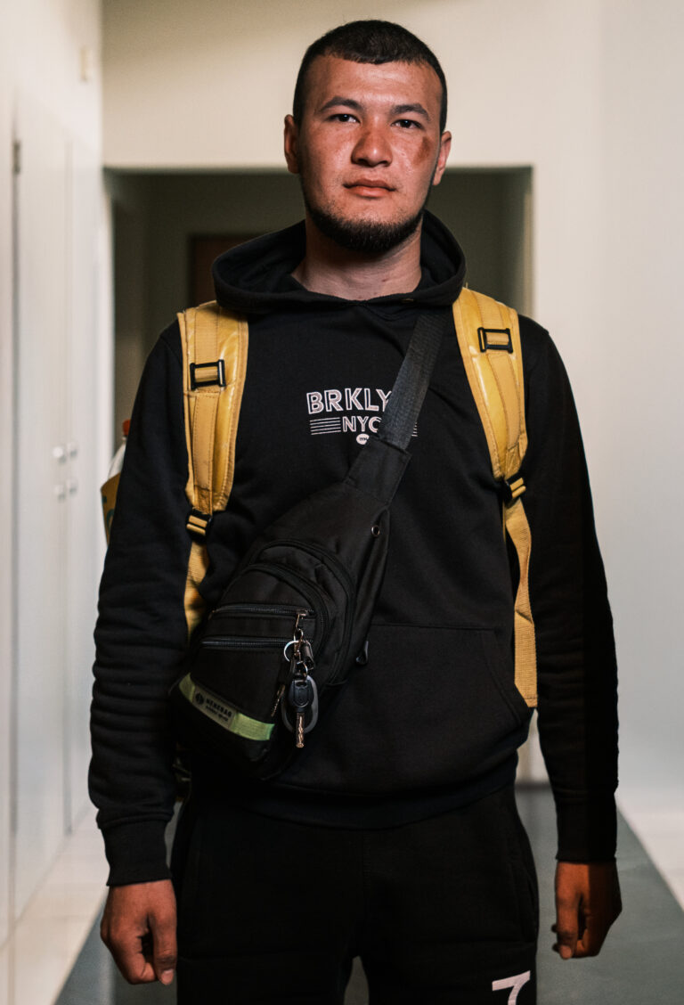 A delivery worker in a black hoodie, with yellow backpack straps and a black crossbody bag, faces the camera in a doorway.