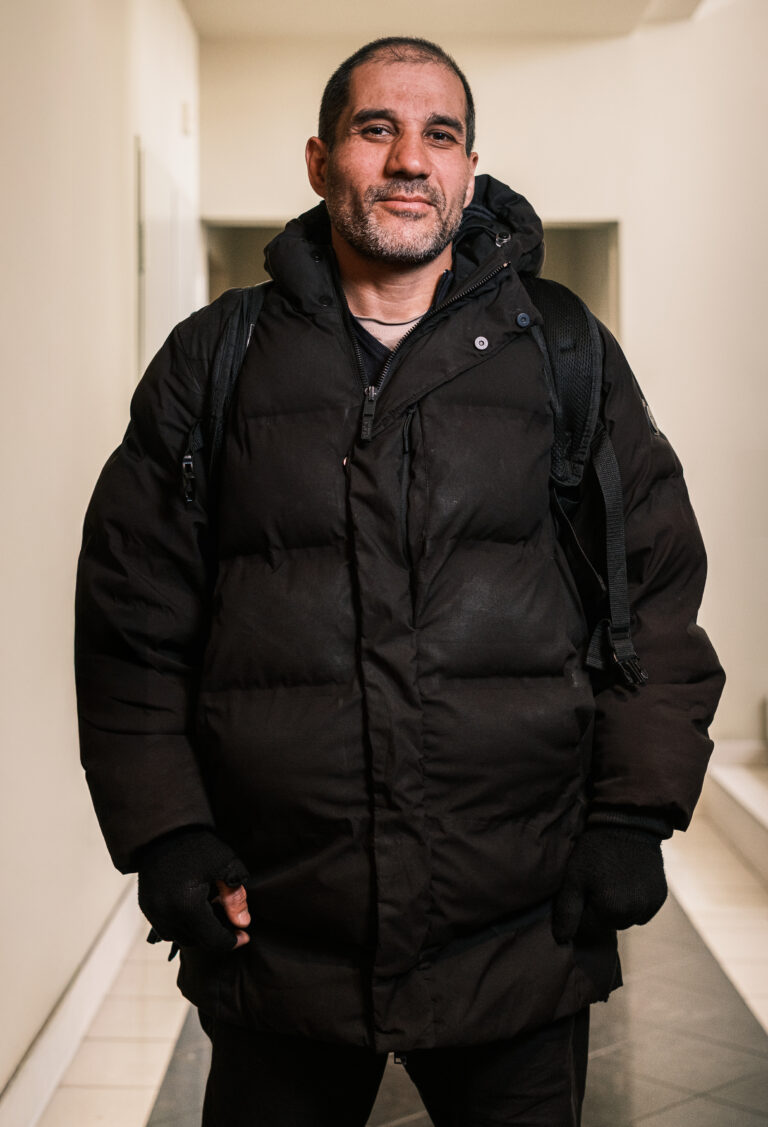 A delivery worker in a black padded jacket stands in a doorway with hands in the jacket pockets.