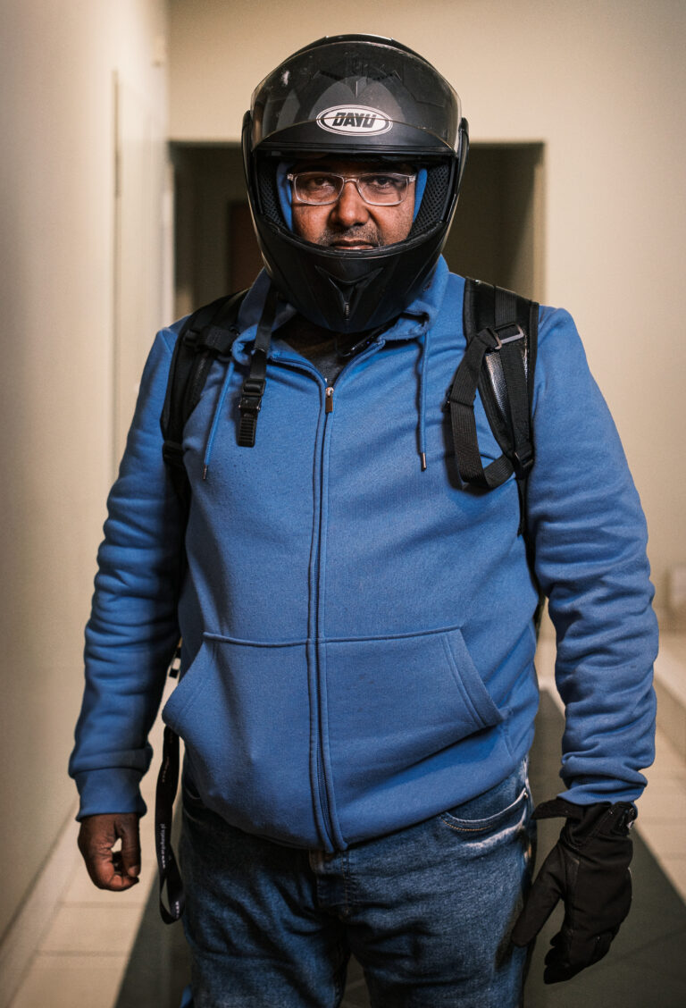A delivery worker in a blue hoodie wears glasses and a black full-face helmet in a doorway.