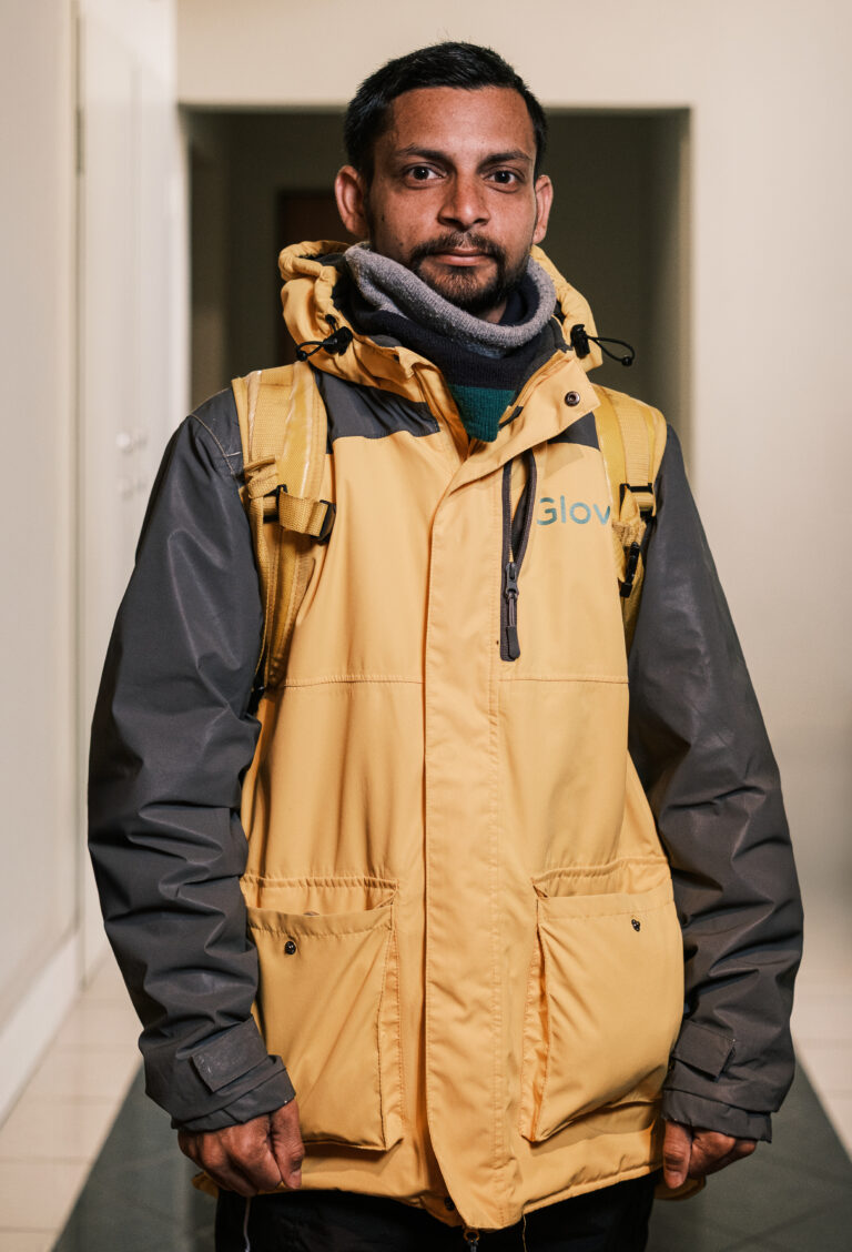 A delivery worker in a yellow and grey jacket and dark scarf stands in a doorway with yellow backpack straps visible.