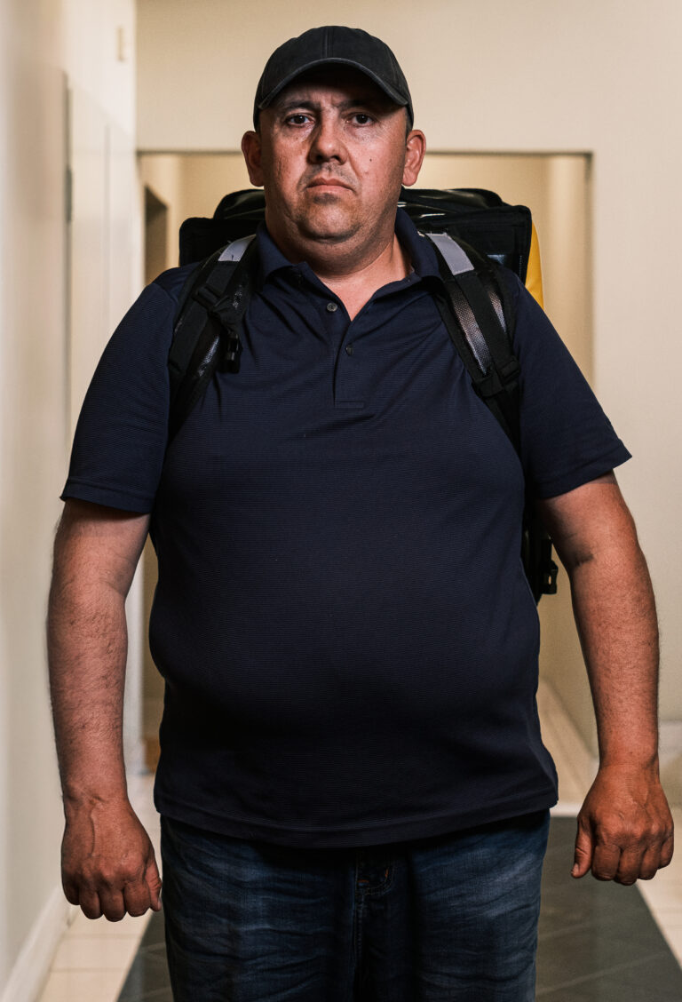 A delivery worker in a navy polo shirt and dark cap stands in a doorway with a backpack.