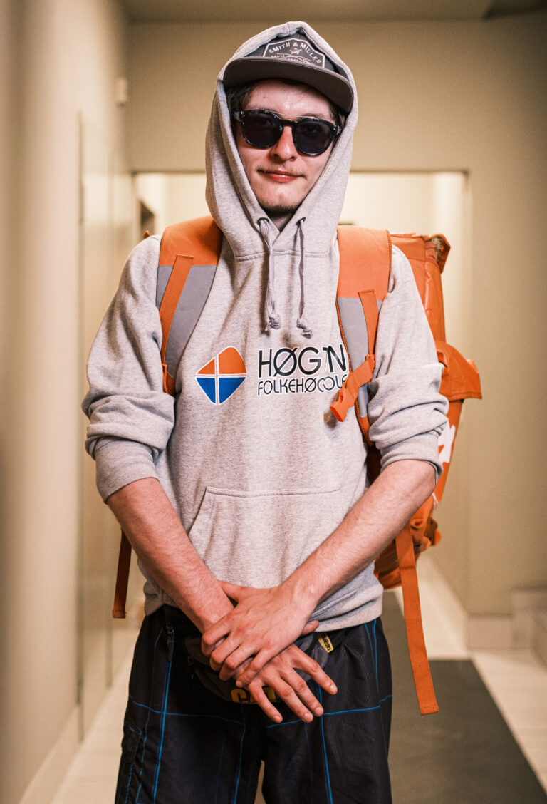A delivery worker in a grey hoodie and sunglasses, with an orange backpack, holds hands together in a doorway.
