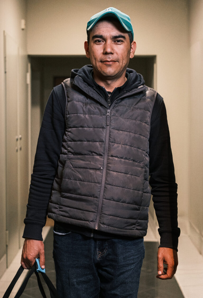 A delivery worker in a grey quilted vest and turquoise cap holds a bag by its handles in a doorway.