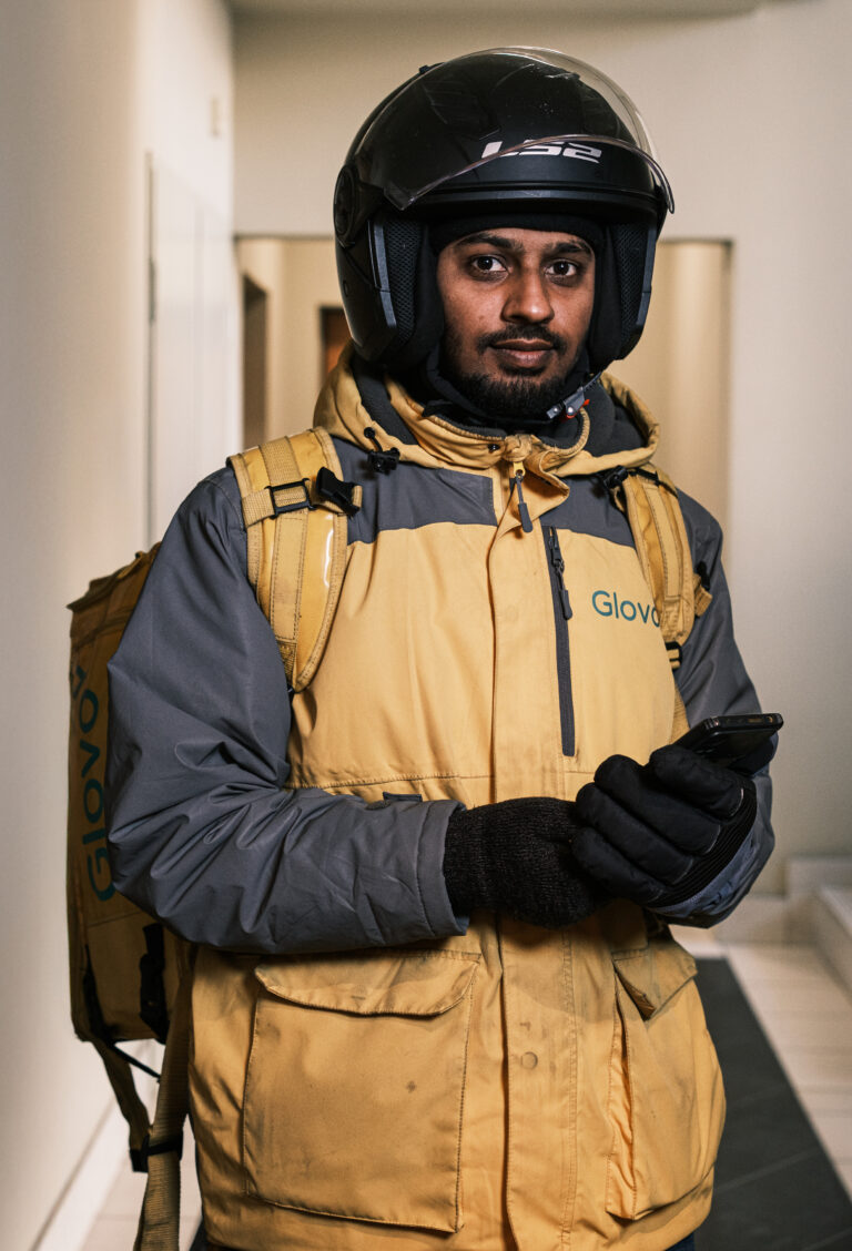 A delivery worker in a yellow and grey jacket and black helmet holds gloved hands together in a doorway.