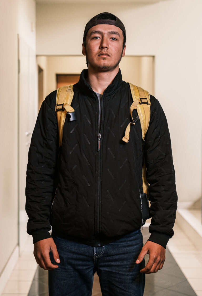 A delivery worker in a black jacket and yellow backpack stands facing the camera in a doorway.