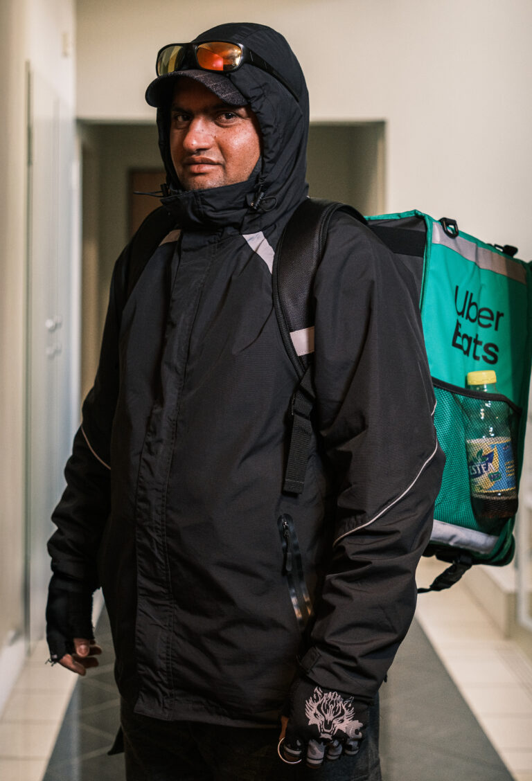 A delivery worker in a black hooded jacket turns slightly sideways beside a green delivery bag in a doorway.