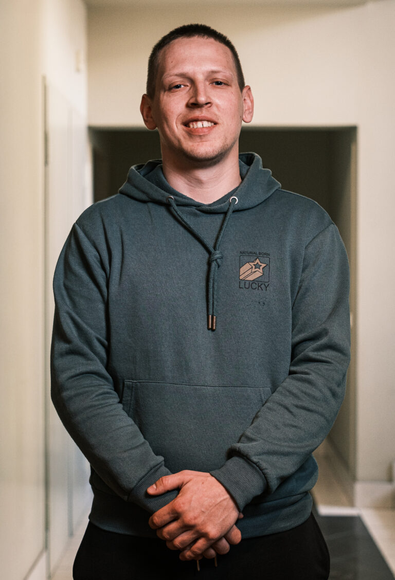 A smiling delivery worker in a dark green hoodie holds hands together at waist level in a doorway.
