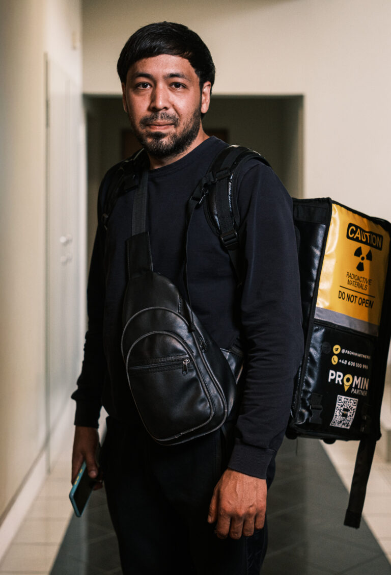 A delivery worker in a black jacket and crossbody bag stands beside a yellow and black delivery bag in a doorway.